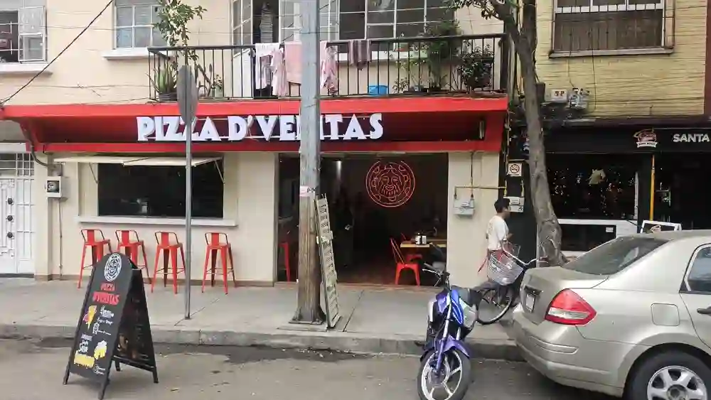 Fachada roja de Pizza D'Veritas de día, con bancos altos y la pizarra en la banqueta