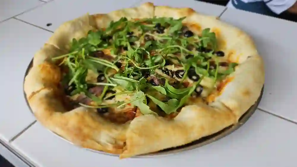 Pizza con arúgula y aceitunas negras, de orilla alta, recién horneada